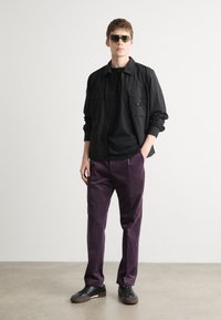 Chemise noire à boutons au-dessus d'un T-shirt noir, assortie à un pantalon violet et des baskets noires. Les accessoires incluent des lunettes de soleil et une bague.