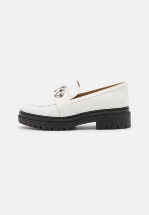 Mocassins en cuir verni blanc avec un accessoire en métal argenté, semelle épaisse en caoutchouc noir et bout arrondi. Texture lisse, design facile à enfiler.