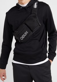 Borsa a tracolla nera in nylon con due tasche con zip e una tracolla regolabile, dotata del logo "HUGO" in bianco. Indossata sopra un felpa nera.