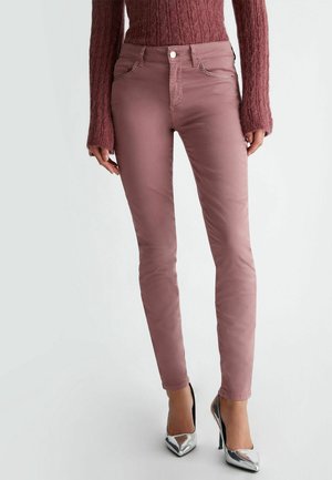 Femme portant un pantalon slim couleur mauve, un pull en maille texturée assorti et des talons hauts argentés pointus, debout devant un fond uni.