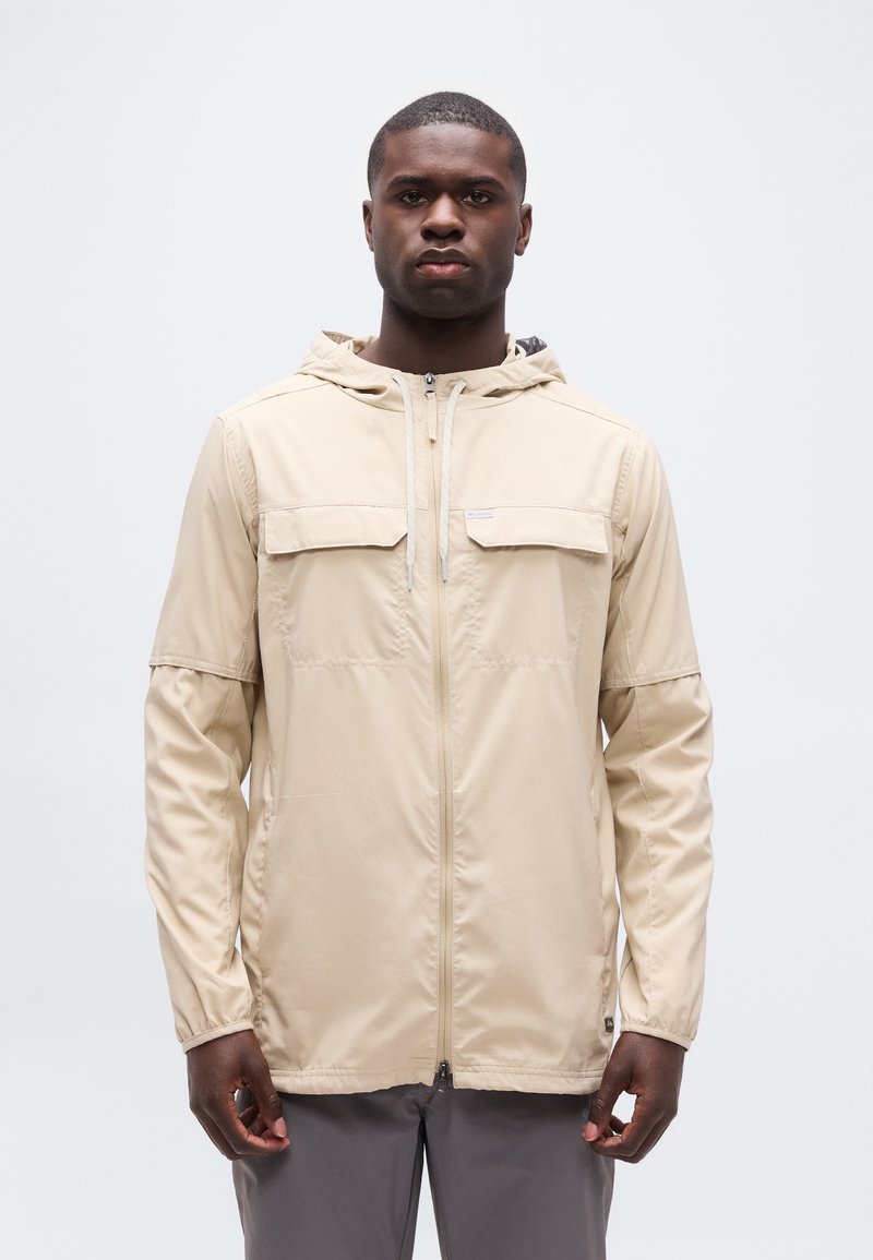 Άνδρας που φοράει μπεζ φούτερ με κουκούλα και φερμουάρ, με τσέπες στο στήθος και γκρι παντελόνι, στέκεται μπροστά σε απλό λευκό φόντο.