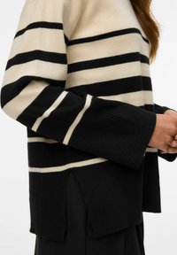 Maglione a righe nere e crema con una vestibilità ampia, polsini a coste, fessure laterali e una texture morbida. Le righe alternano larghezza.