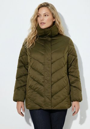 Femme aux longs cheveux blonds portant une doudoune matelassée vert olive et un pantalon foncé, debout devant un fond clair uni.