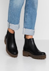 Botas de piel negra hasta el tobillo con paneles laterales elásticos, suela de plataforma marrón gruesa, punta redonda y detalles de costura. Llevadas con jeans azules arremangados.