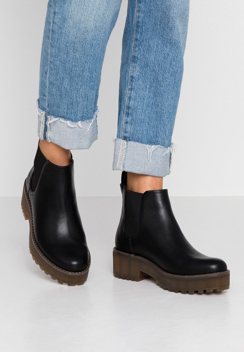 Botas de piel negra hasta el tobillo con paneles laterales elásticos, suela de plataforma marrón gruesa, punta redonda y detalles de costura. Llevadas con jeans azules arremangados.