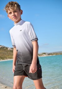 Kortärmad rashguard med gradient från vitt till svart med prickmönster, kombinerad med svarta badshorts. Bärs av en pojke på en strand.