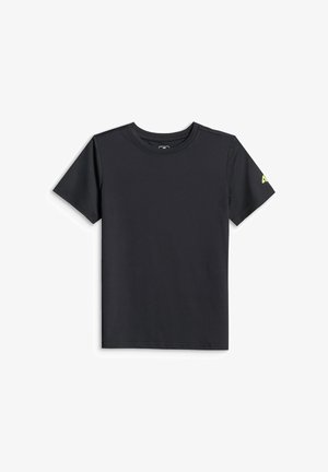 Garçon aux cheveux mi-longs portant un t-shirt noir, un short gris à motifs, des chaussettes blanches avec des rayures noires et des baskets noir et blanc, debout sur un fond gris.