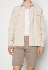 Camisa chaqueta de tela beige con frente de botones, cuello puntiagudo, dos bolsillos en el pecho y mangas largas, combinada con pantalones cortos marrón claro.