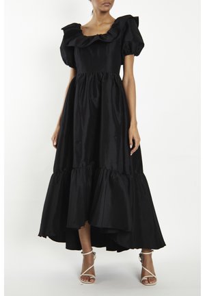 Femme portant une robe noire à manches bouffantes avec un col carré à volants et un ourlet asymétrique, associée à des sandales blanches à lanières.