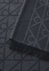 Sort vævet tekstil med et mønsterdesign, der indeholder geometriske former. Frynsede kanter og varierende teksturer skaber visuel interesse.