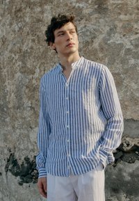 Young man in blue and white vertical striped shirt and white pants standing against textured weathered wall, looking to the side.