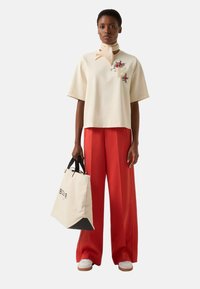 Béžový top s krátkými rukávy a květinovou výšivkou, zkombinovaný se širokými červenými kalhotami. V ruce drží béžovou tašku se černými detaily a logem. Bílé tenisky.