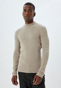 Beige ribbel gebreide trui met een hoge hals en lange mouwen, gecombineerd met zwarte broek. De textuur van de trui is zacht en aansluitend.