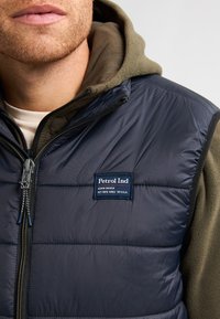 Marineblau gesteppte Weste mit Frontreißverschluss, graugrüner Kapuzenpullover darunter und "Petrol Ind"-Logopatch. Verfügt über eine glatte Textur und kontrastierende Farben.