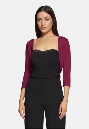 Donna con capelli lisci e scuri indossa un top nero senza spalline con cintura e un cardigan corto rosa scuro, in piedi davanti a uno sfondo uniforme.