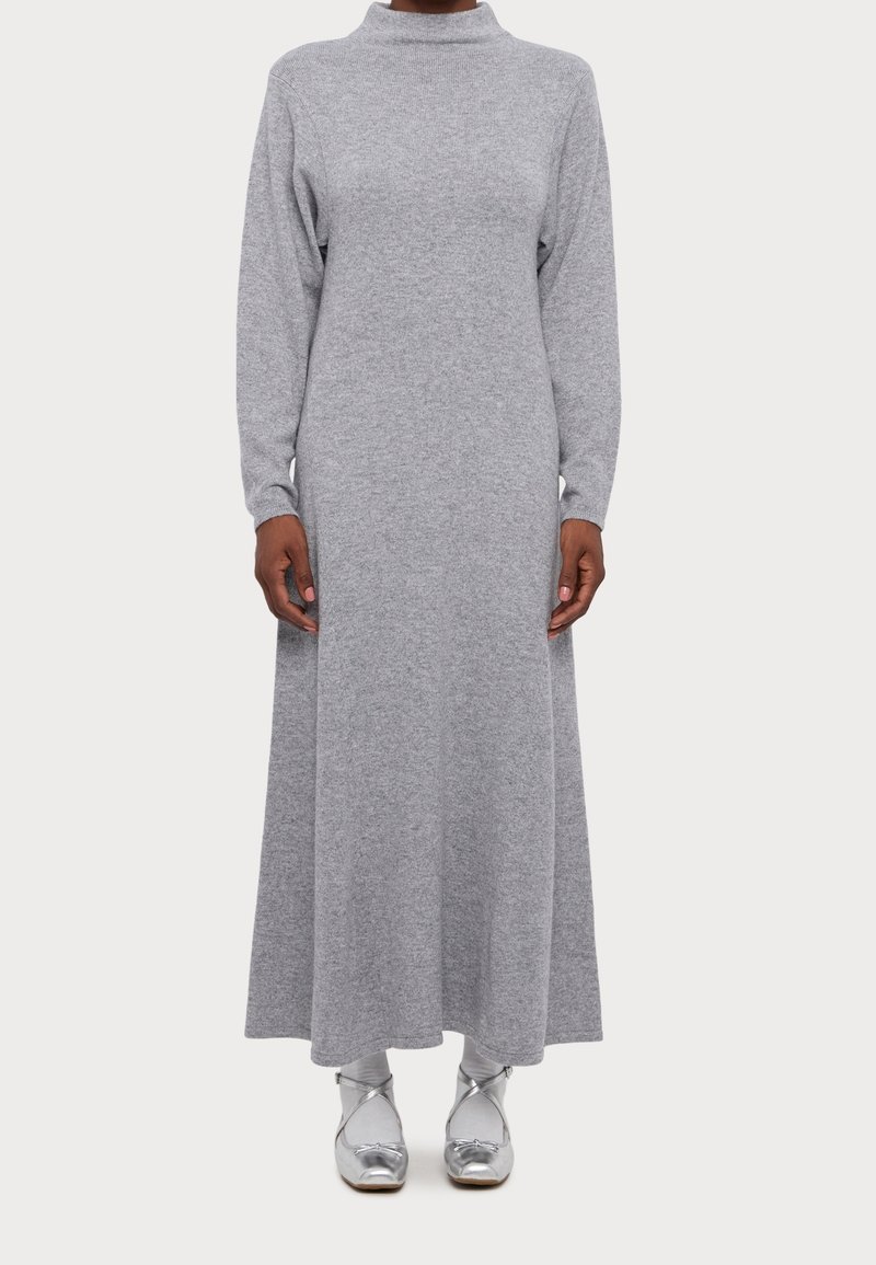 Robe longue grise en maille avec un col montant et des manches longues, présentant une coupe ample et arrivant aux chevilles. Portée avec des chaussures argentées.