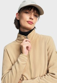Veste en polaire beige avec un col montant et une fermeture éclair ; texture douce ; associée à une casquette de couleur claire arborant un logo et un col roulé noir en dessous.