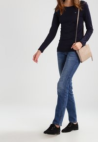 Marineblauw overhemd met lange mouwen, lichtblauwe slim-fit jeans en zwarte veterschoenen. Accessoires zijn een beige crossbody tas.