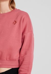 Mujer vistiendo un sudadera corta de color rosa polvo con dobladillo acanalado y un logotipo de estrella y chevron bordado en el pecho.