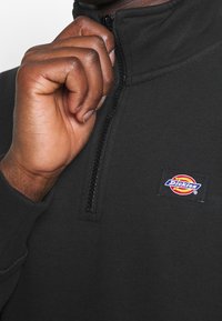 Pull-over noir à demi-fermeture éclair, en tissu doux, présentant une surface texturée et un petit logo coloré « Dickies » sur le côté supérieur gauche.