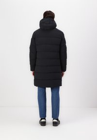 Doudoune noire avec capuche, présentant un design matelassé et des coutures verticales. Associée à un jean bleu et des chaussures noires.