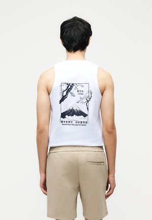 Man die met zijn rug naar voren staat, draagt een wit mouwloos shirt met een zwart-witte afbeelding van de Mount Fuji met bomen en Japanse tekst, beige korte broek.