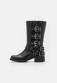 Black leather boot with a tall shaft, featuring multiple silver buckles and straps, a circular accent, and a chunky heel. Matte finish.