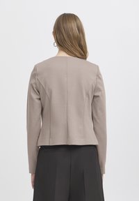 Femme aux cheveux châtain clair portant un blazer beige et un pantalon noir, vue de dos sur un fond clair uni.