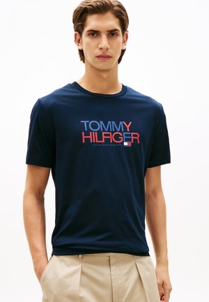 Jeune homme aux cheveux châtain clair portant un t-shirt bleu marine Tommy Hilfiger et un pantalon beige, debout devant un fond blanc uni.