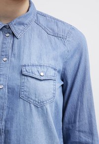 Ljusblå denimskjorta med klassisk krage, tryckknappsstängning och två bröstfickor. Tyget har en mjuk, något strukturerad yta.