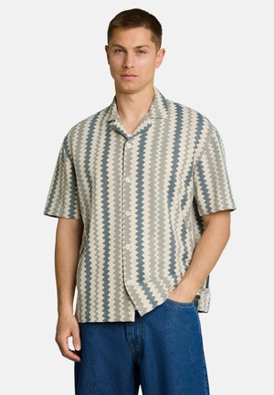 Mann trägt kurzärmliges Hemd mit vertikalen Zickzackstreifen in Beige und Blau sowie blaue Jeans, steht mit einer Hand in der Tasche.