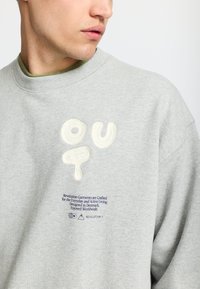Lichtgrijze sweatshirt met getextureerde, crèmekleurige lettering "OU" en grafische details. Inclusief blauwe tekst aan de onderkant. Zachte katoenstof.