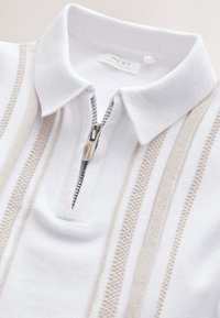 Biela polokošeľa s čiastočným zipsom, zdobená béžovými textúrovanými pruhmi a rebrovaným golierom. Materiál je hladký a mierne elastický.
