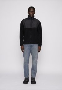 Sort fleecejakke med et kontrasterende gråt vandtæt øverste panel, lynlås lukning, høj krave og sidelommer, båret med lyseblå jeans.