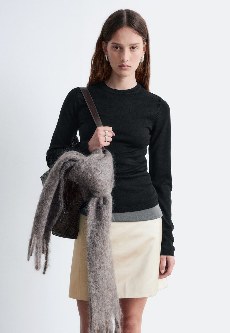 Schwarzer, langärmeliger Pullover über einem grauen Shirt, kombiniert mit einem beigen Rock. Hält einen grauen, flauschigen Schal und eine geflochtene braune Tasche.