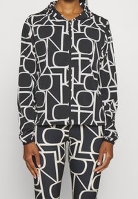 Schwarze Jacke mit weißem geometrischem Muster, mit Reißverschluss und Kapuze. Aus leichtem Stoff gefertigt, kombiniert mit passenden Leggings im gleichen Muster.