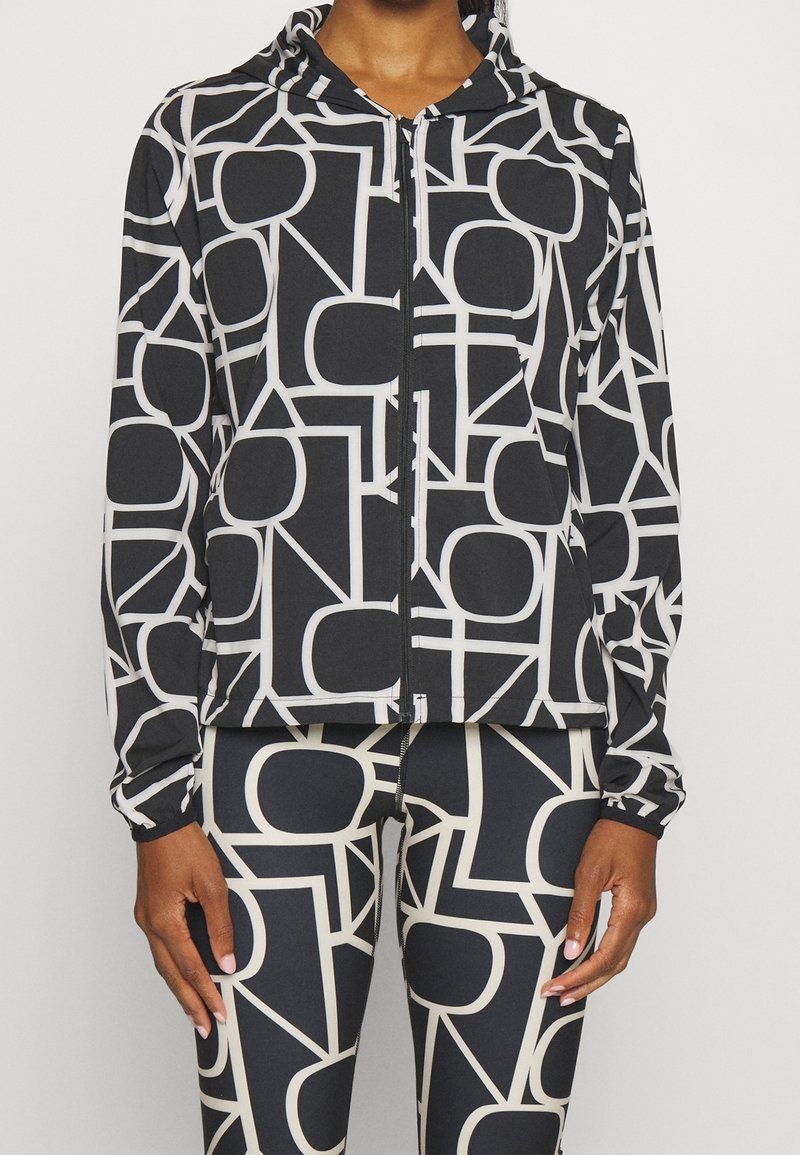 Schwarze Jacke mit weißem geometrischem Muster, mit Reißverschluss und Kapuze. Aus leichtem Stoff gefertigt, kombiniert mit passenden Leggings im gleichen Muster.