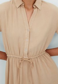 Beżowa sukienka zapinana na guziki, z kołnierzykiem, elastycznym paskiem w talii i wiązaniem z przodu. Gładki materiał o subtelnym połysku i krótkich rękawach.