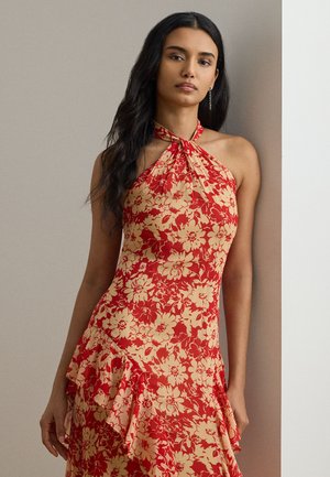 Žena s dlhými tmavými vlasmi, oblečená v červeno-béžových kvetinových šatách bez rukávov s volánovými detailmi, stojaca na neutrálnom pozadí.