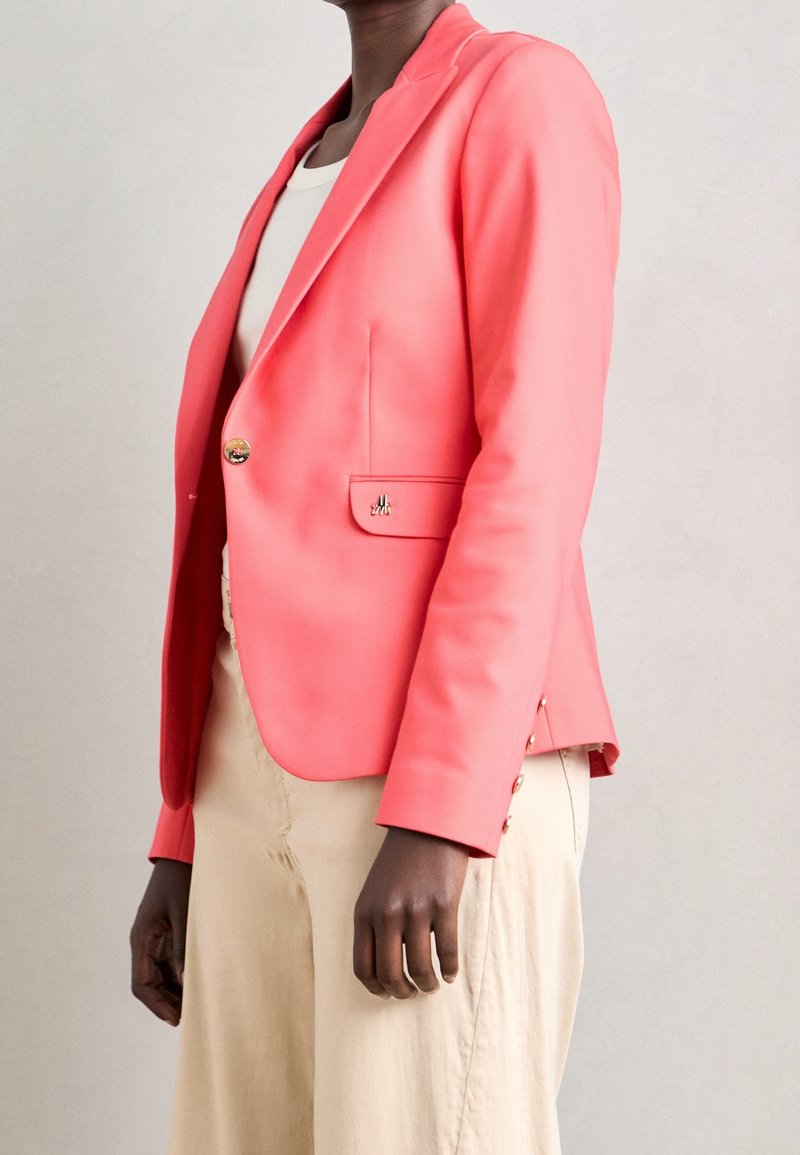 Blazer corail avec une fermeture à un seul bouton et un accent doré. Il présente une coupe ajustée, des revers et des détails à boutons sur les manches.