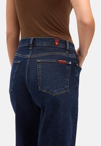 Ciemnoniebieskie dżinsy z denimu z wysokim stanem, wyposażone w dwie tylne kieszenie, subtelne detale przeszyć oraz czerwoną metkę z logo na pasku w talii.