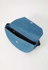 Borsa crossbody in denim blu con base nera, forma rotonda, chiusura magnetica e tasca con zip interna. Texture liscia, design elegante.