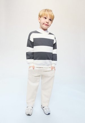 Plavo dijete stoji sa rukama u džepovima, nosi crno-bijeli prugasti džemper, krem hlače i sive tenisice na bijeloj pozadini.