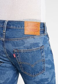 Siniset farkut, joissa on maastokuvio, varustettuna ruskealla nahkapatchilla, jossa on kohokuvioitu logo, ja tavallisilla takataskuilla, joissa on ompeluyksityiskohtia.