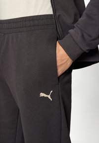 Μαύρο αθλητικό παντελόνι με κομψό σχεδι ασμό, ελαστική μέση, πλαϊνές τσέπες και μπεζ λογότυπο Puma στο αριστερό μηρό. Μαλακό, υφασμάτινο ύφασμα με υφή.