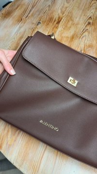 Καφέ δερμάτινη τσάντα χειρός με υφή, χρυσά μεταλλικά στοιχεία και κλείσιμο με καπάκι. Διαθέτει ανάγλυφο λογότυπο "VALENTINO" στο εμπρόσθιο μέρος.