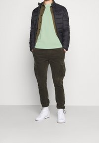 Puffer bunda v černé barvě, pod ní olivově zelená košile, světle zelené tričko, tmavé cargo kalhoty a bílé tenisky. Neformální vrstvený outfit.