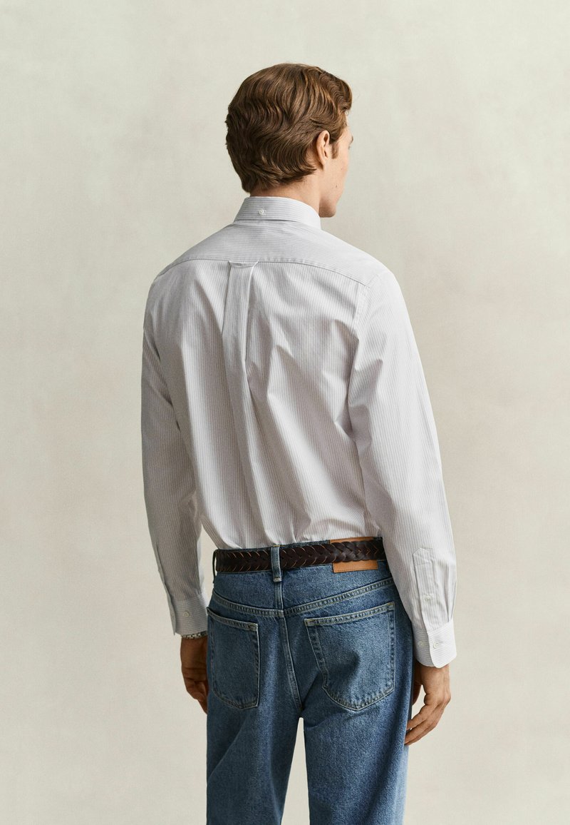Man seen from behind wearing a white long-sleeve shirt and blue jeans with a brown braided belt against a plain background.
