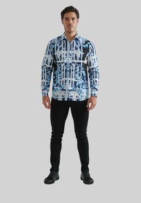 Chemise à manches longues bleu et noir, avec un motif graphique comportant des éléments architecturaux. Portée avec un pantalon slim noir et des chaussures noires.
