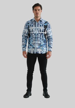 Chemise à manches longues bleu et noir, avec un motif graphique comportant des éléments architecturaux. Portée avec un pantalon slim noir et des chaussures noires.